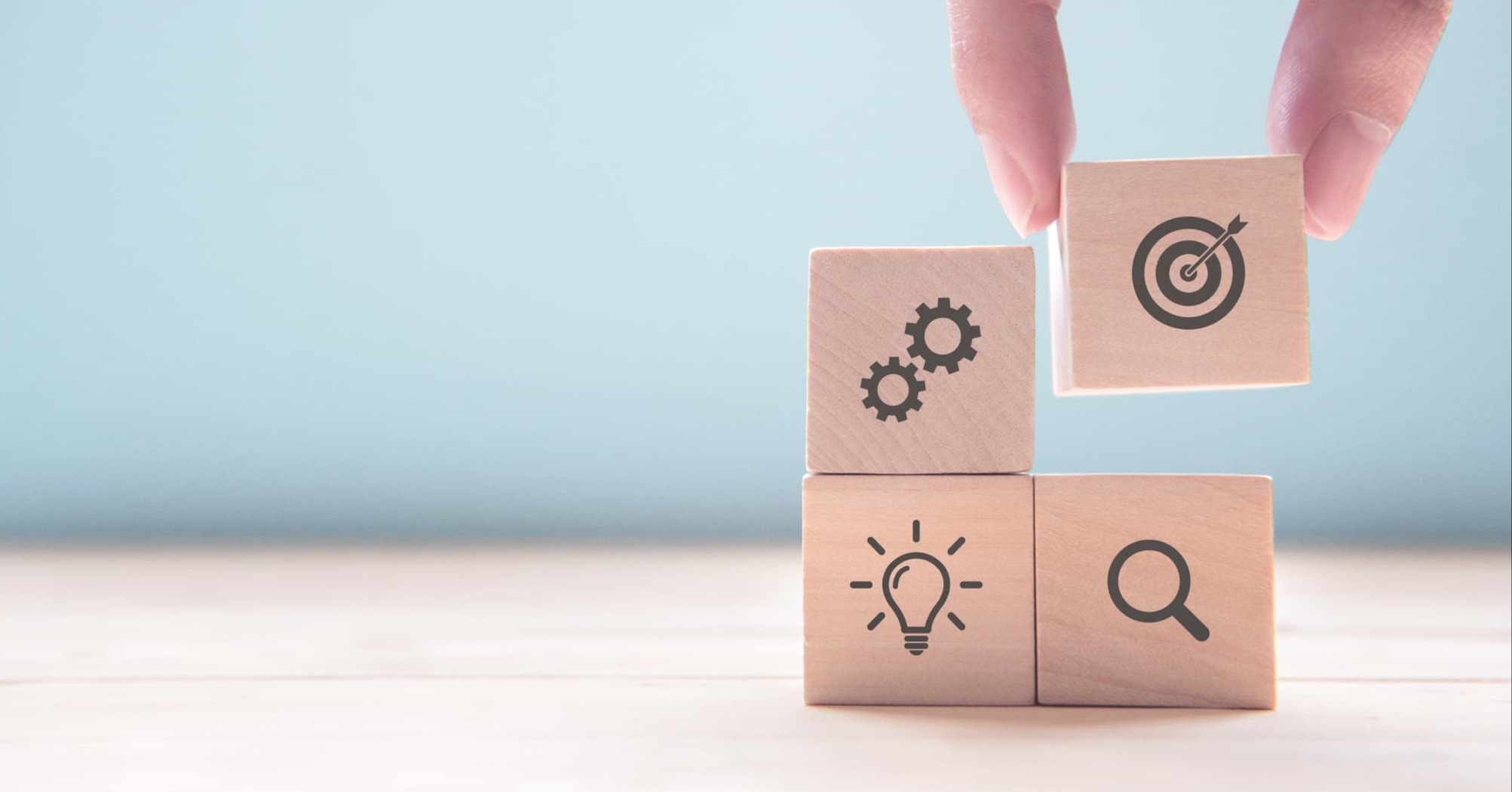 a close-up of someone stacking toy blocks, with different symbols representing different business goals: gears, target, lightbulb, magnifying glass.