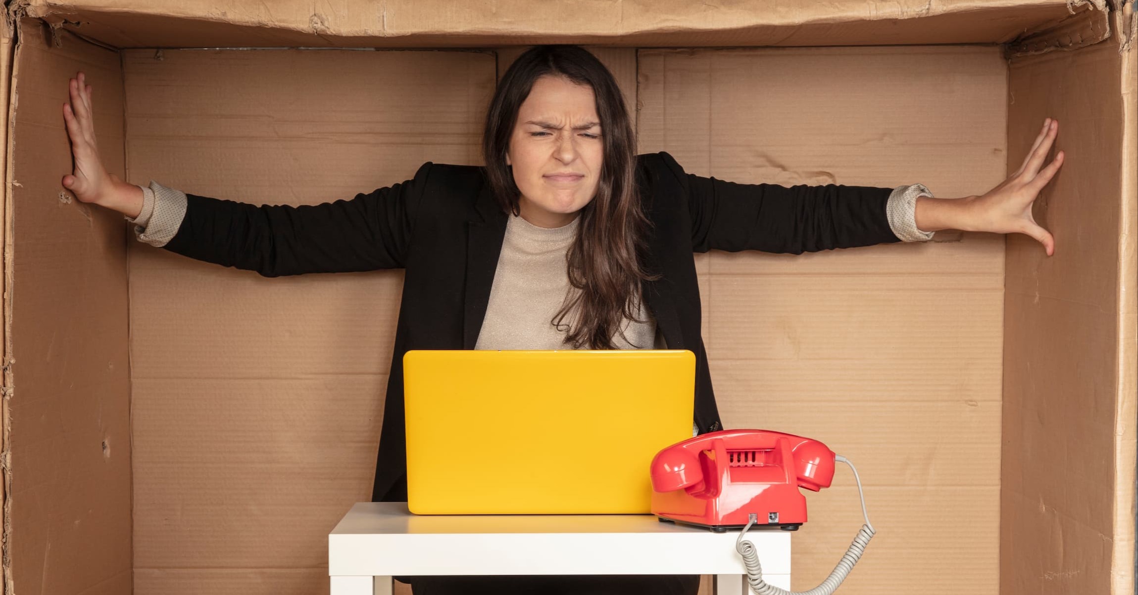 a worker sitting at a small desk in a cardboard box, pushing against the edges. representing being put in a box or having limited resources at work.